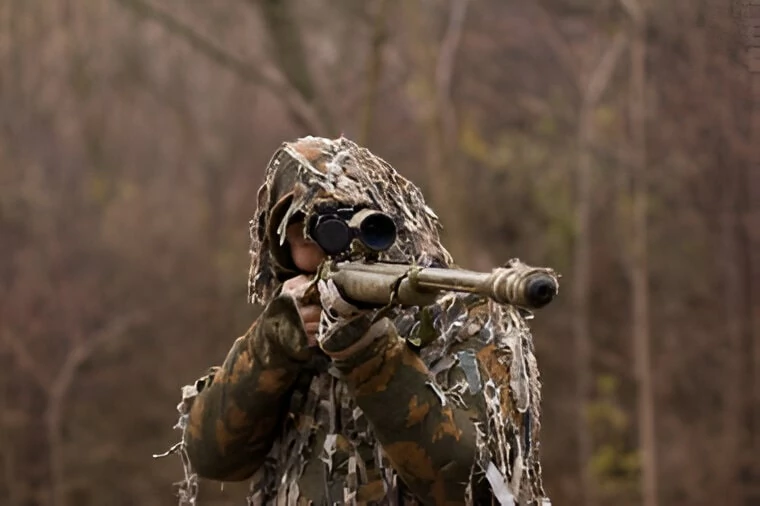 A man dressed in camouflage gear is aiming a rifle, focused and prepared for action in a natural environment.